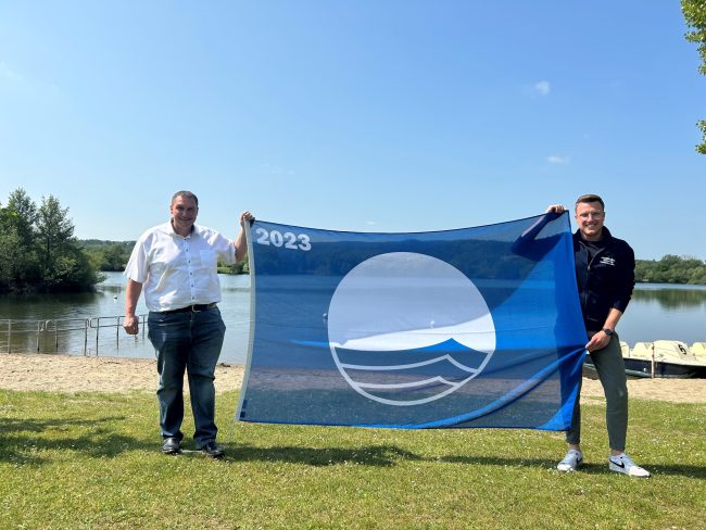 Bürgermeister Jochen Weber und Yannic Wildner (Gemeinde Michelau i.OFr.) mit der Blauen Flagge am Rudufersee bei Schwürbitz. Das Geländer im Hintergrund hilft Menschen, die nicht so gut zu Fuß sind, sicherer ins Wasser zu kommen. Foto: Gemeinde Michelau i. OFr. Bürgermeister Jochen Weber und Yannic Wildner (Gemeinde Michelau i.OFr.) mit der Blauen Flagge am Rudufersee bei Schwürbitz. Foto: Gemeinde Michelau i. OFr.