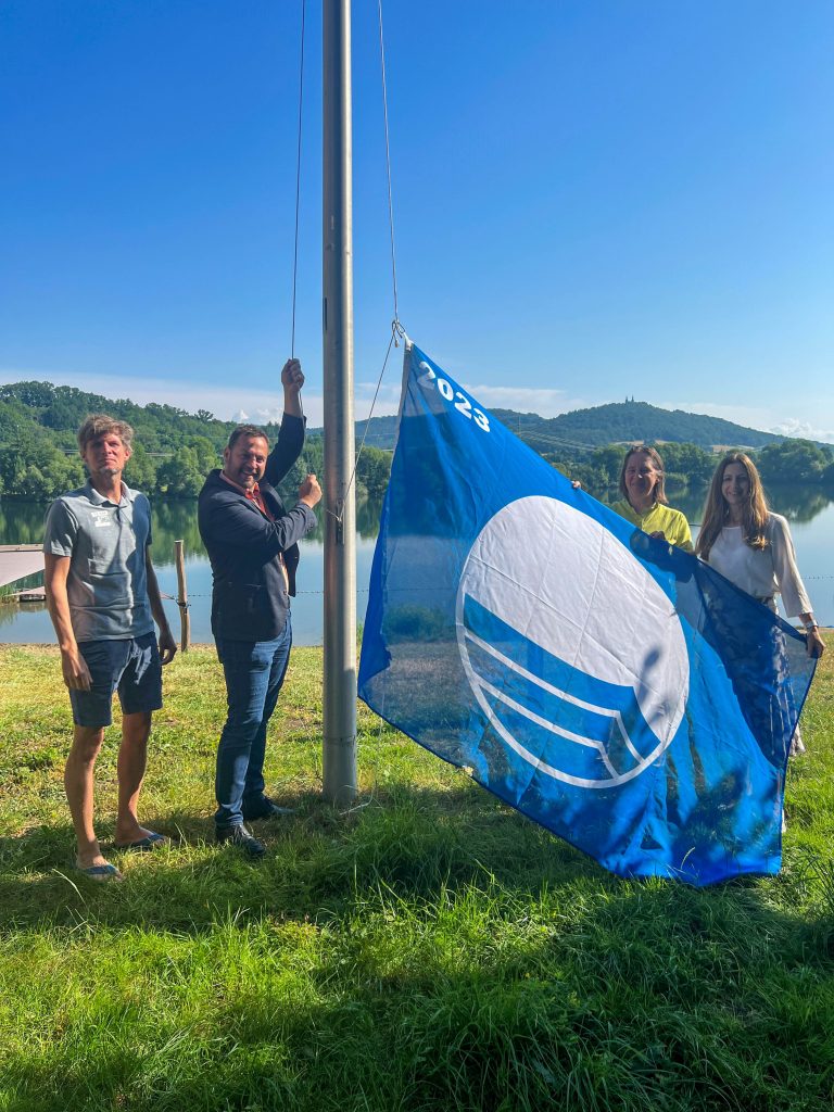 Martin Lüders(Freizeit GmbH), Bürgermeister Mario Schönwald, Anne Schmitt (Flussparadies Franken) und Anne Schneider (Tourismusleiterin Bad Staffelstein) hissen gemeinsam die Blaue Flagge 2023 am Bad Staffelsteiner Ostsee. Foto: Freizeit GmbH